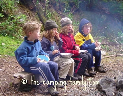Four small children sitting on a low timber bench by a campfire, without any table