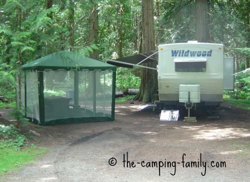 A screen tent erected next to an RV in a woodland setting